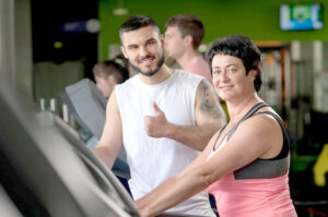 Young male fitness coach with middle aged female client on treadmill machine. Attractive brunette woman working out in gym with personal trainer. Healthy lifestyle concept. Selective focus